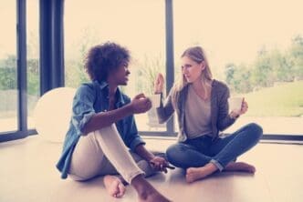 Multiethnic women sit on the floor, laughing and drinking coffee.