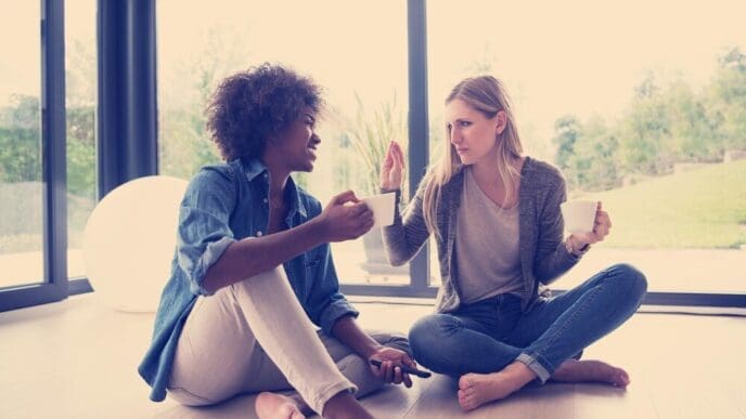 Multiethnic women sit on the floor, laughing and drinking coffee.