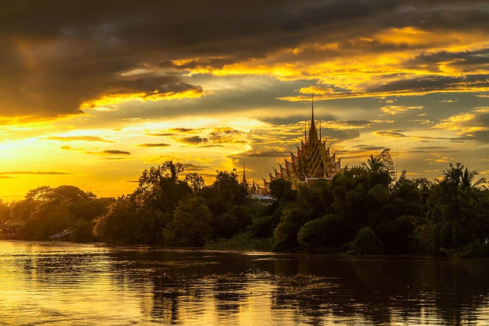 A serene evening scene shows a temple complex reflected in the Nan River at Raft Village in Phitsanulok, Thailand.