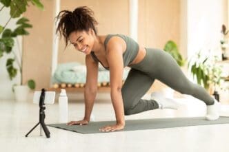 An African American woman in workout clothes does mountain climbers on a yoga mat while looking at a smartphone.
