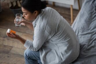 Overhead shot shows a multiracial woman in a bedroom looking sadly at pills while holding a glass of water.