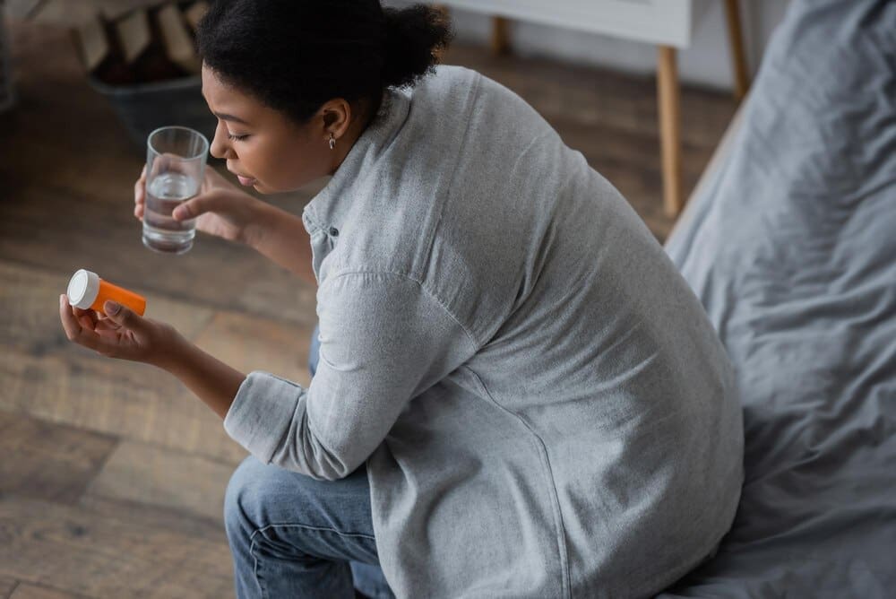 Overhead shot shows a multiracial woman in a bedroom looking sadly at pills while holding a glass of water.