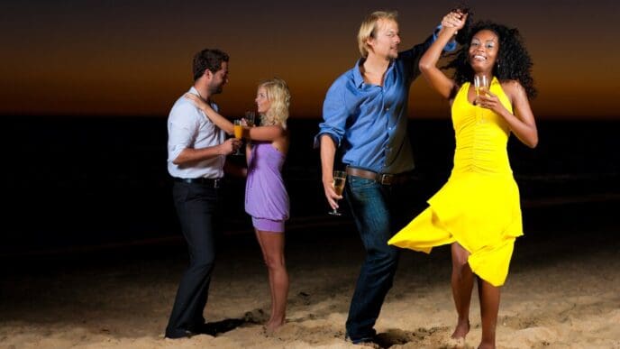 People are dancing and celebrating on a sandy beach, likely at a party.