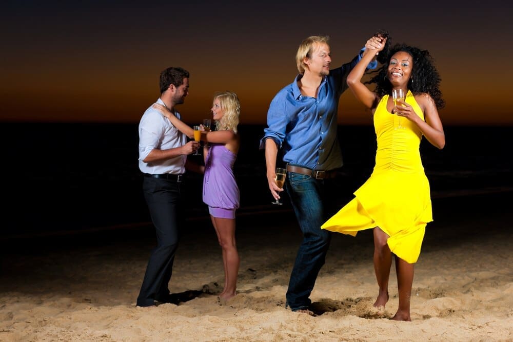 People are dancing and celebrating on a sandy beach, likely at a party.