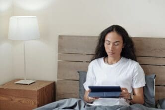 A young man sits in bed, looking at a tablet computer with a thoughtful expression.