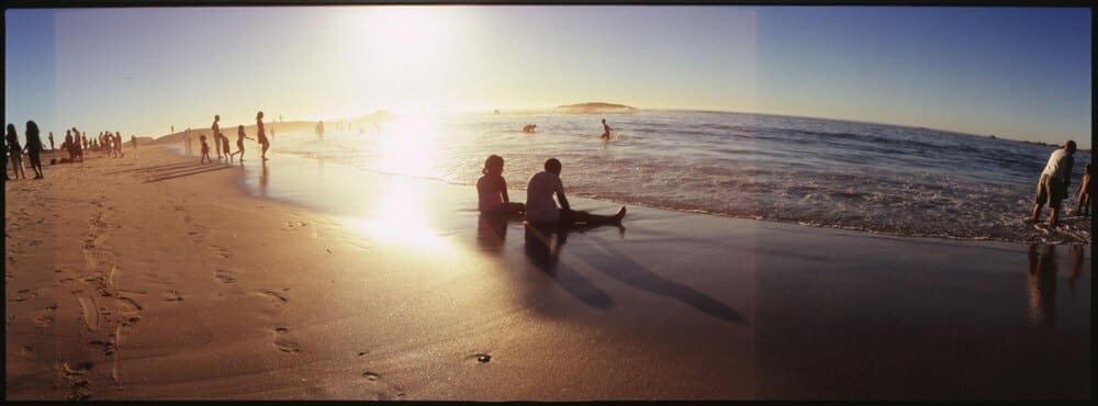 People of various ages stand and sit on a sandy beach.
