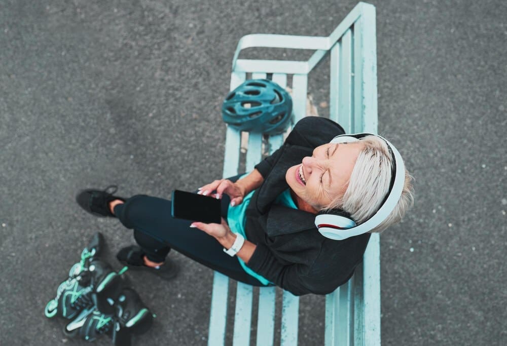 A mature woman wearing roller skates prepares to skate while listening to a podcast on her phone, smiling.