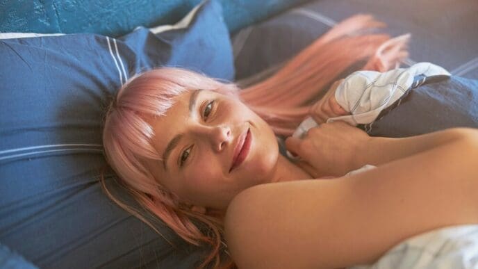 Attractive young woman with pink hair awakens in a large, comfortable bed.
