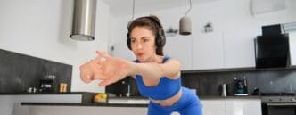 A fit woman stretches her body while watching a gym workout tutorial on a device at home.