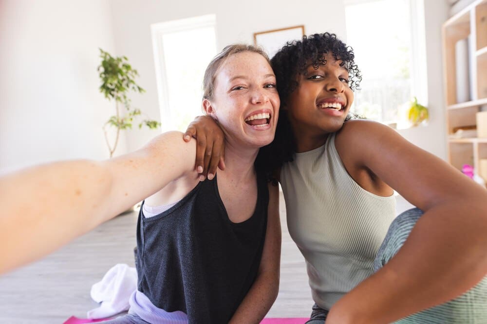 Smiling diverse female friends in athletic wear take a selfie together indoors.