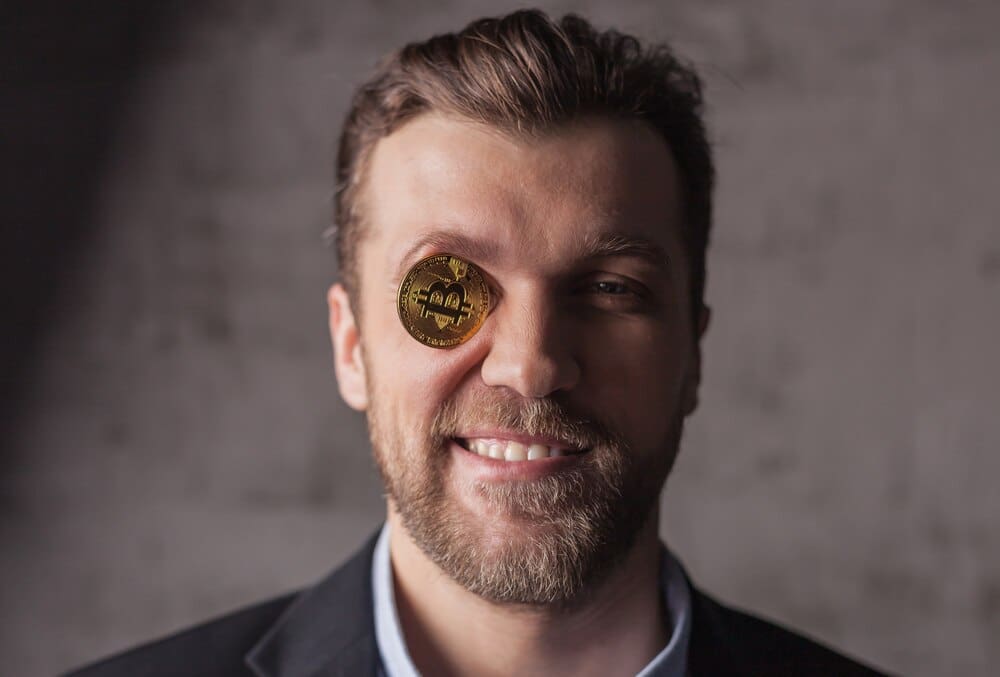 A man with a smiling expression has a Bitcoin logo in place of one of his eyes.