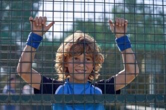 A young boy with curly hair holds onto a metal tennis net, looking away toward the sunlight on a sports field.