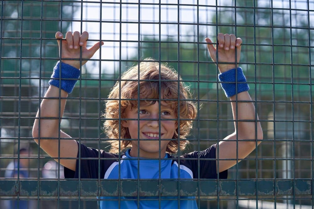A young boy with curly hair holds onto a metal tennis net, looking away toward the sunlight on a sports field.