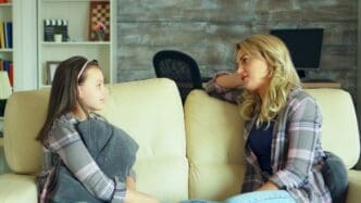 A young girl sits on a couch in a living room and converses with her mother.
