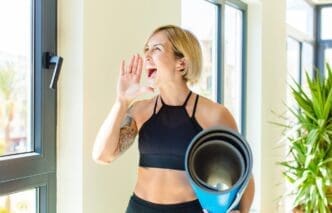 A blonde woman with her mouth open wide, yelling with an angry expression, and her hand beside her mouth.