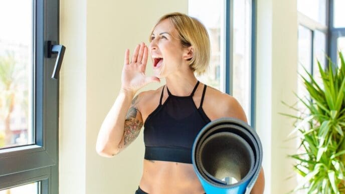 A blonde woman with her mouth open wide, yelling with an angry expression, and her hand beside her mouth.