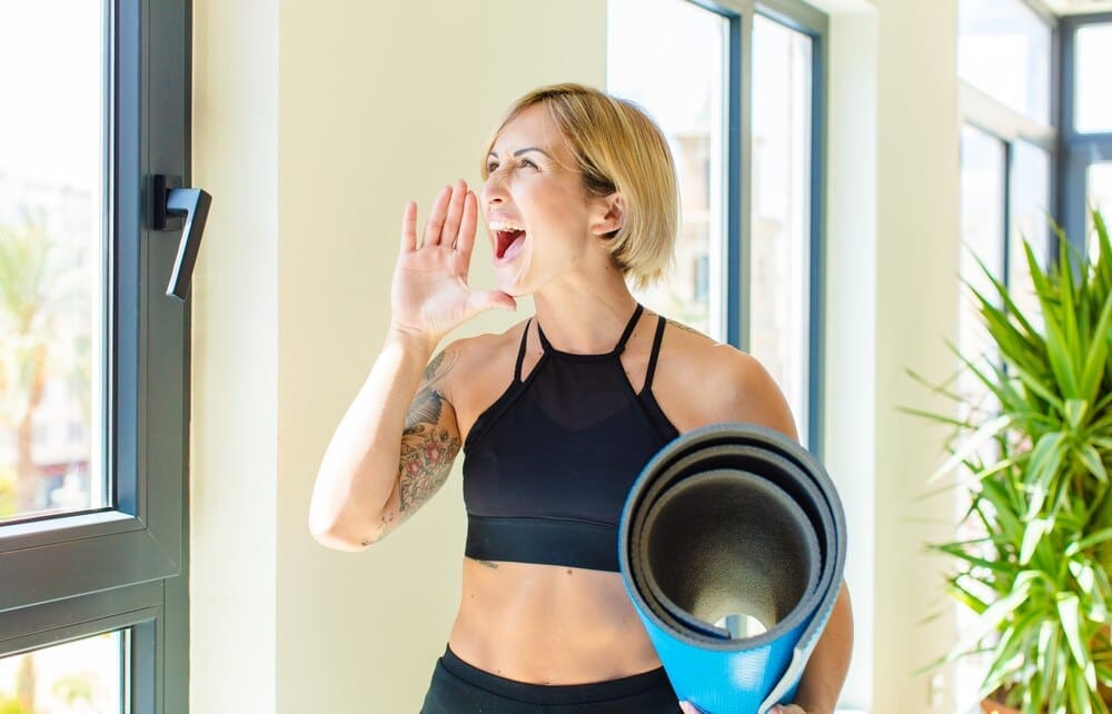 A blonde woman with her mouth open wide, yelling with an angry expression, and her hand beside her mouth.