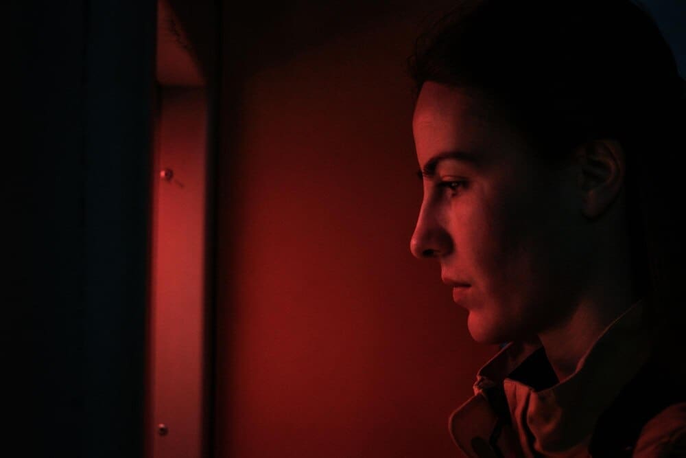 Profile of a young woman standing in a dark room, illuminated by an unseen light source.