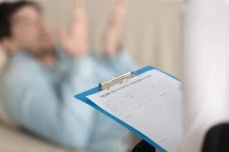 A close-up on a clipboard displaying a "Medical Patient Information" form, held by a blurred medical professional, with a patient lying on a couch in the background during a consultation.