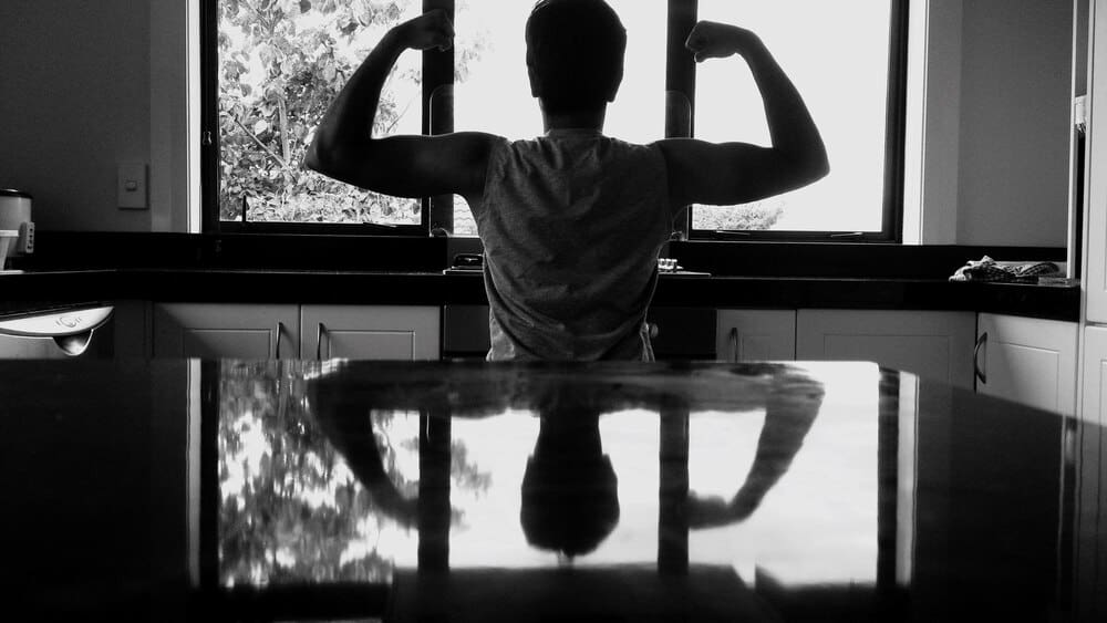 A man flexes his muscles in a kitchen, viewed from the back.