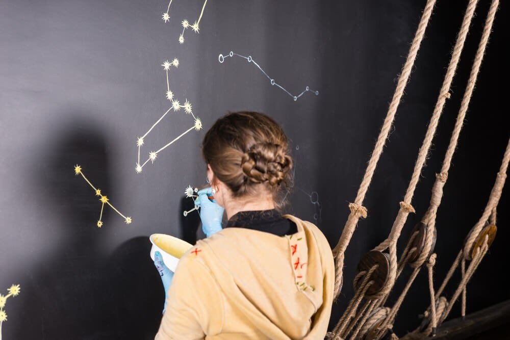 Rear view of a woman painting star constellations on a black chalkboard within a room designed to resemble a ship's deck.