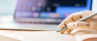 A robotic hand holds a pencil near a notebook and laptop, suggesting artificial intelligence or automation in writing.