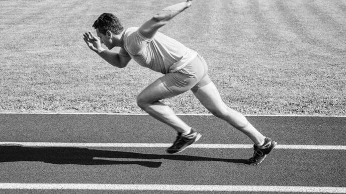 A runner bursts forward from the starting position on a stadium track on a sunny day, illustrating the beginning of a sprint race.