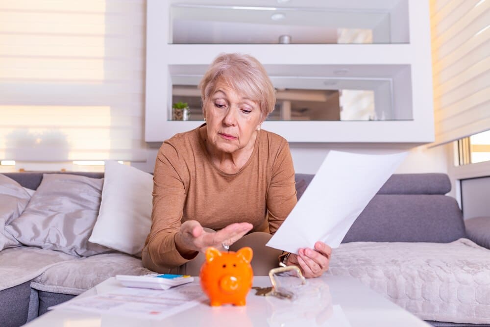 A senior woman puts a coin into a piggy bank while calculating the money inside.
