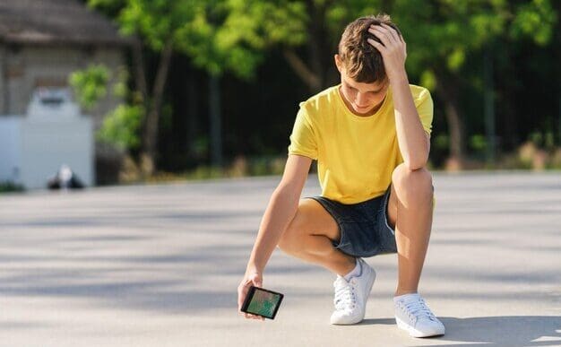 A young boy with a sad expression clutches a broken cell phone while standing in a skatepark.