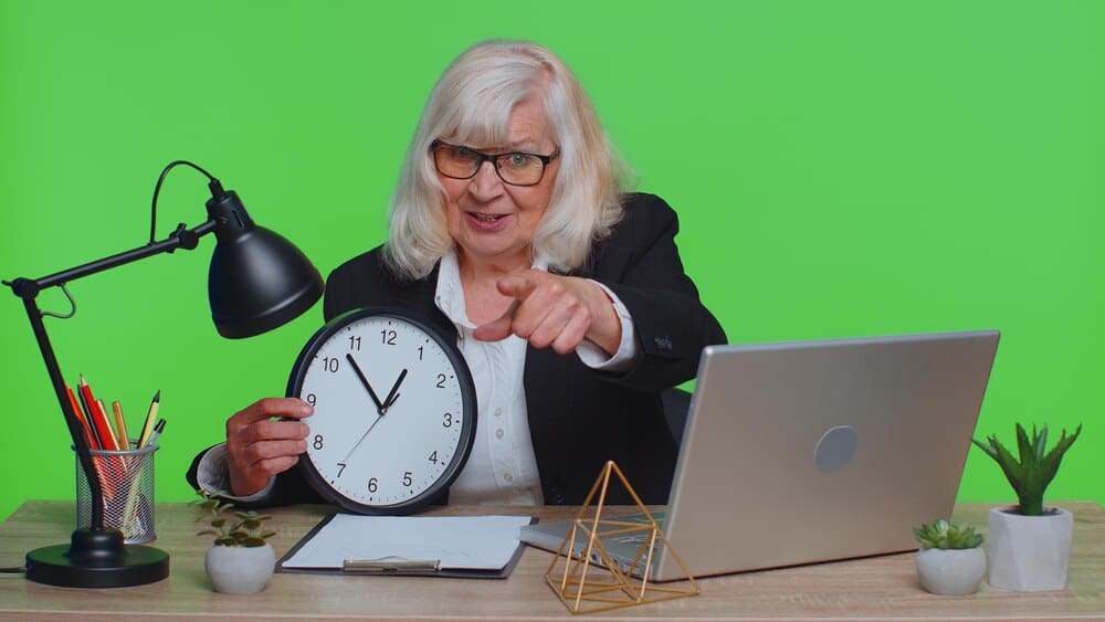 A senior businesswoman smiles and gives a thumbs-up while pointing to a clock on the wall.