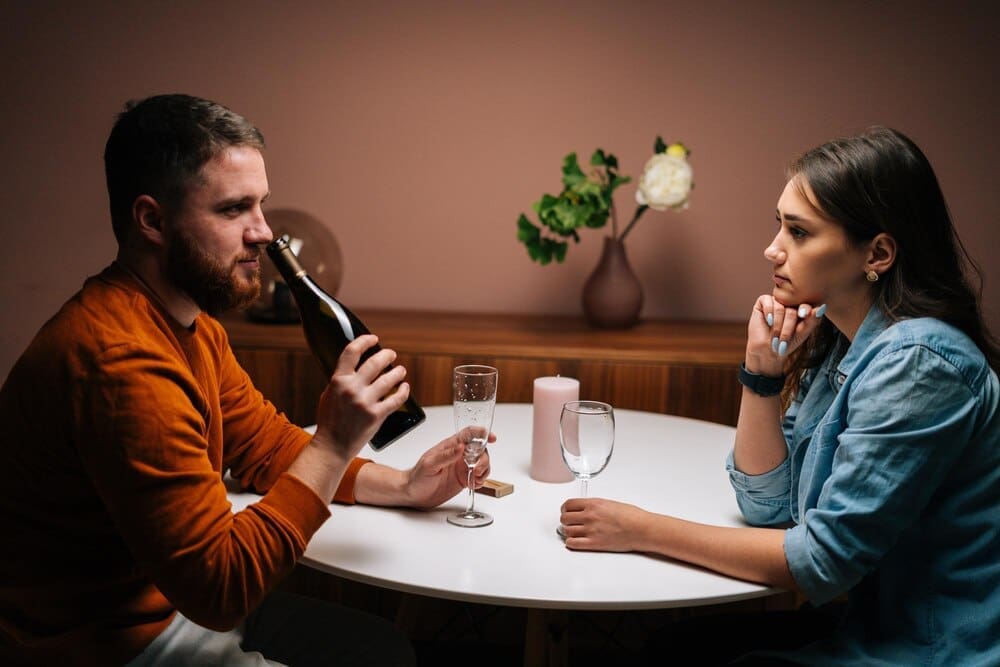 A young man with a beard sniffs a bottle of wine while talking to his girlfriend at a table during a romantic dinner.