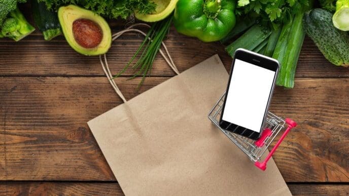 Smartphone displaying a food ordering app next to a shopping trolley and fresh green vegetables on a wooden table.