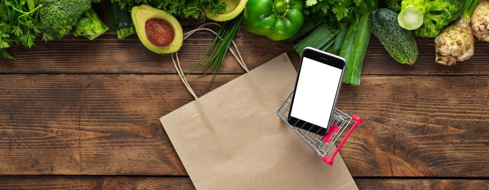 Smartphone displaying a food ordering app next to a shopping trolley and fresh green vegetables on a wooden table.