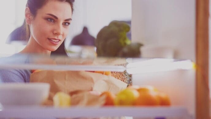A smiling woman reaches into a refrigerator to grab fresh fruit, illustrating a healthy eating concept.
