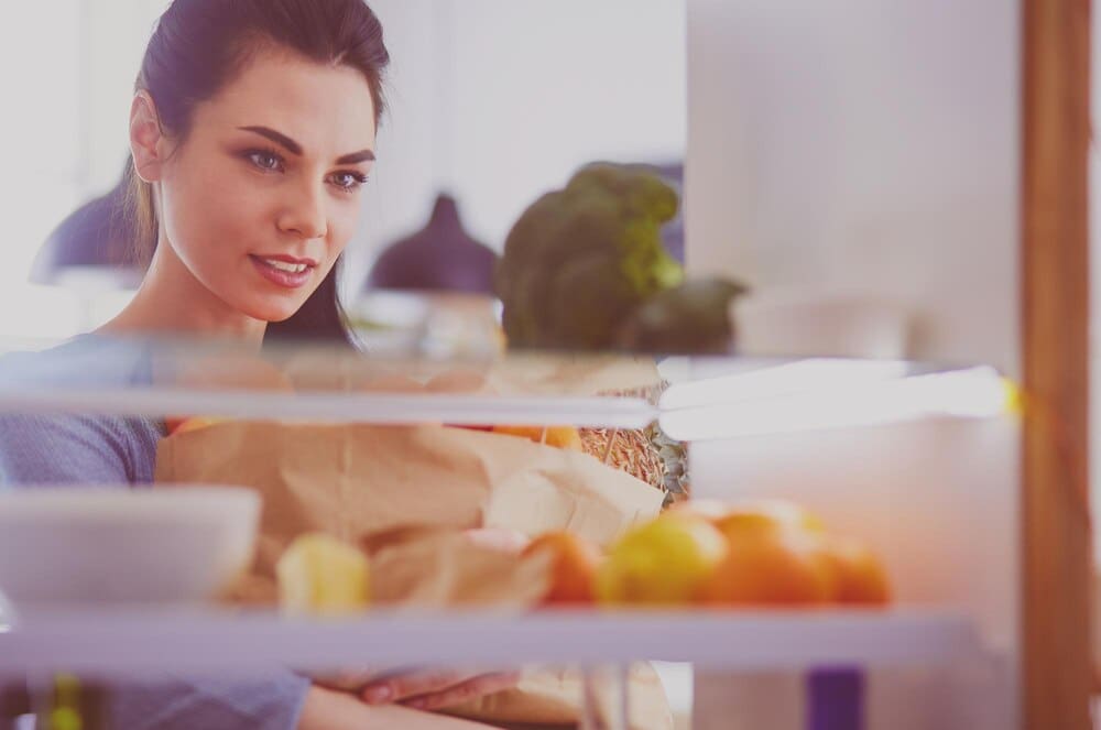A smiling woman reaches into a refrigerator to grab fresh fruit, illustrating a healthy eating concept.