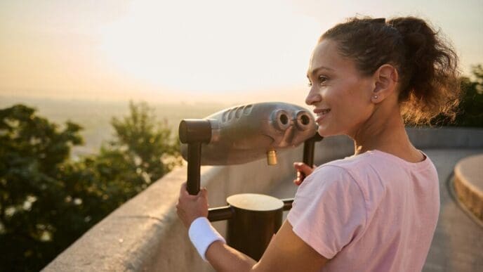 A woman in a pink t-shirt looks through a telescope at a city skyline.
