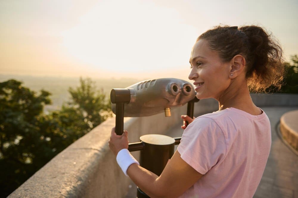 A woman in a pink t-shirt looks through a telescope at a city skyline.