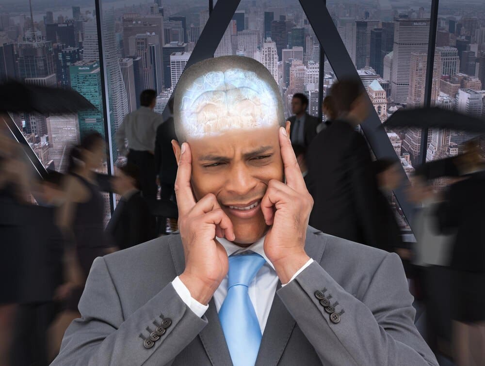 A stressed businessman puts his fingers to his temples in an office with a large window overlooking a city.