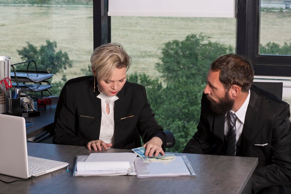 A Caucasian businesswoman and a bearded businessman in a black suit count money together in a modern office.