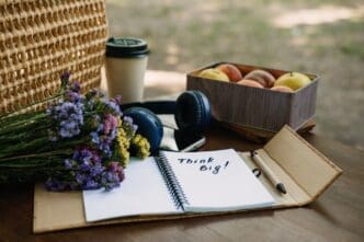 A partially visible hand writes "Think Big" in an open notebook on a wooden table, outdoors.