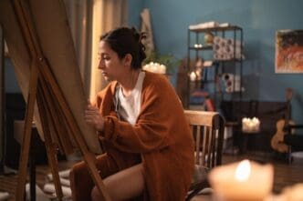 A young girl sits in a chair at home, focused on sketching or drawing on a canvas at an easel.