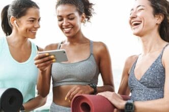Three young women in athletic wear smile while standing on a beach and holding fitness mats, with one using a mobile phone.
