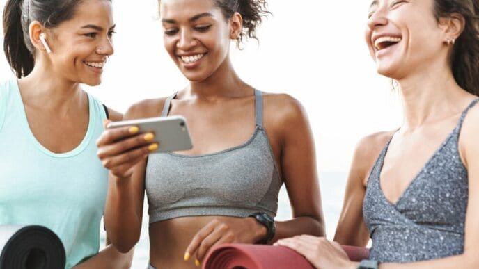Three young women in athletic wear smile while standing on a beach and holding fitness mats, with one using a mobile phone.