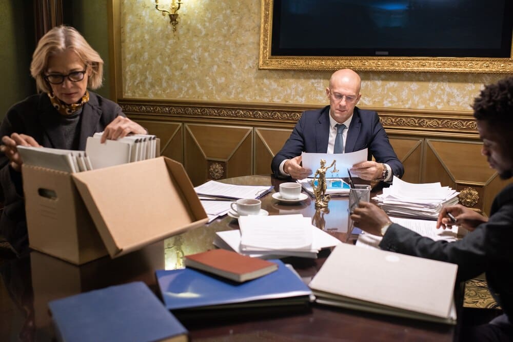 Three lawyers, dressed in modern attire, collaborate around a table covered in documents.