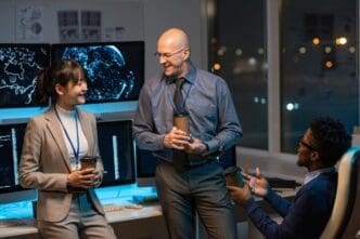 Three coworkers smile and chat over drinks while seated at a desk with computer monitors.