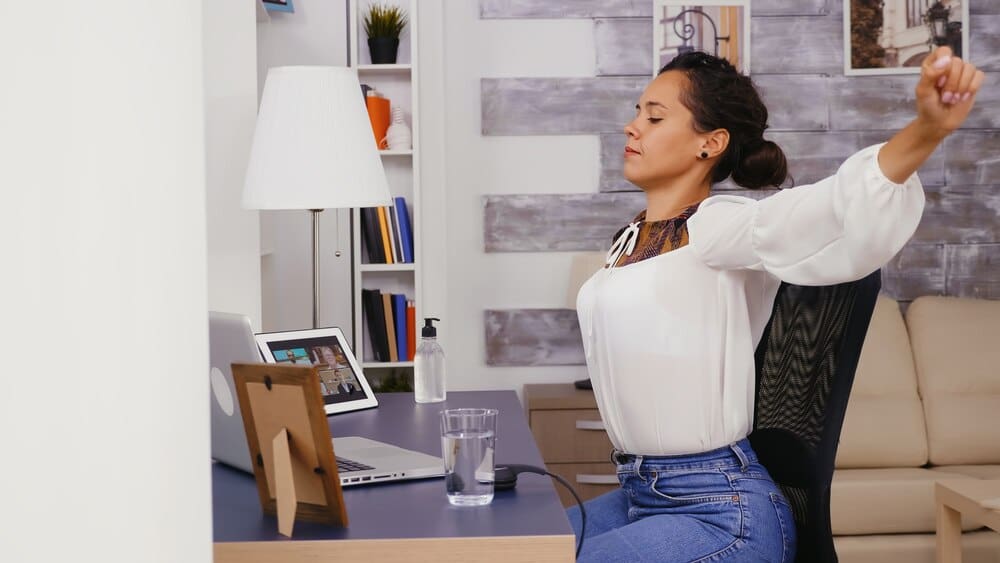 A woman in a home office stretches her back, appearing tired.
