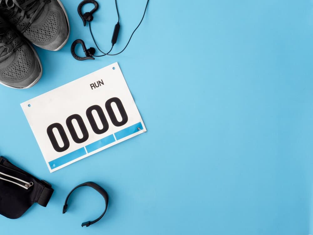 Top-down view of running shoes, a bib number, and running accessories arranged on a blue background, representing a running event.