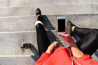 Top-down view of a young woman in athletic wear drinking water while listening to music on her phone.