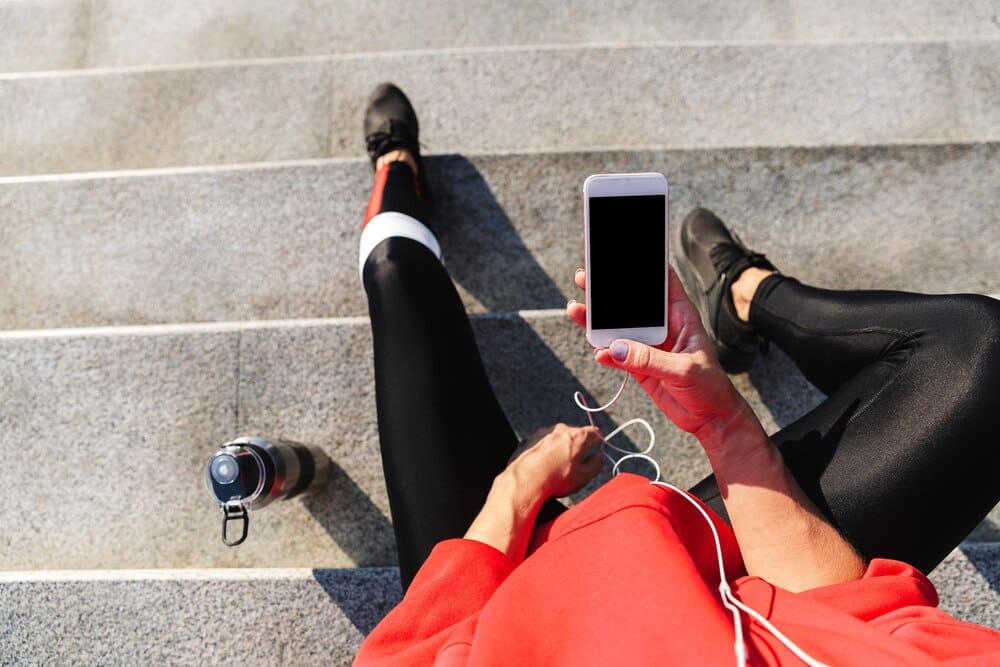 Top-down view of a young woman in athletic wear drinking water while listening to music on her phone.