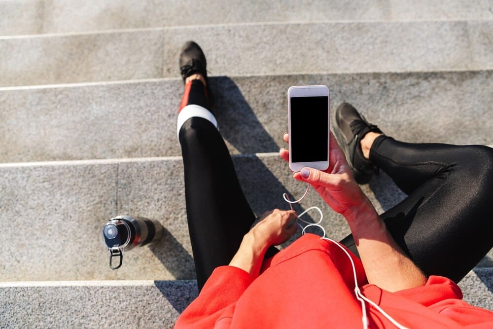 Top-down view of a young woman in athletic wear drinking water while listening to music on her phone.